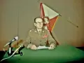 Generał w mundurze, z flagą Polski za plecami, przemawia na konferencji prasowej. To ważny moment dla Wojskowej Rady Ocalenia Narodowego.