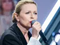 Anna Maria Jopek, w czarnej marynarce, śpiewa do mikrofonu. W tle rozmyte postacie i światła sceny.