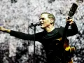 Bryan Adams na koncercie, śpiewa do mikrofonu i gra na gitarze. Wskazuje palcem na publiczność, tworząc niezapomniane chwile.