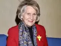 Uśmiechnięta Irena Santor w czerwonej marynarce i wzorzystym szalu, z żółtą rozetką na klapie.