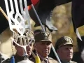Trzech starszych mężczyzn w wojskowych czapkach, trzymających flagi i symbol "upa".