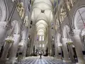 Wnętrze katedry Notre Dame z imponującymi łukami, sklepieniem i żyrandolami. Na szachownicowej posadzce widać pojedynczą postać.