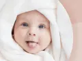 Urocze dziecko z niebieskimi oczami wystawia język, otulone białym kocykiem. Luiza znaczenie imienia - symbol niewinności i radości.
