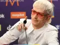 Joost Klein w białej koszuli i różowych okularach przeciwsłonecznych, z charakterystyczną fryzurą, wskazuje palcem w stronę mikrofonu.