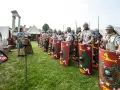 Rzymscy legioniści w pełnym uzbrojeniu, z tarczami i włóczniami, gotowi do bitwy. W tle namioty i ruiny.