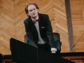 Piotr Ryszard Pawlak w eleganckim garniturze pochyla się nad fortepianem, gotowy do wykonania utworu.