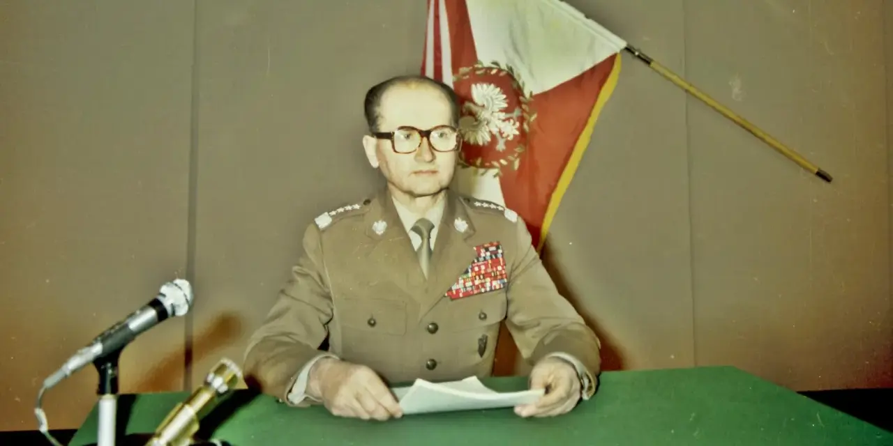 Generał w mundurze, z flagą Polski za plecami, przemawia na konferencji prasowej. To ważny moment dla Wojskowej Rady Ocalenia Narodowego.