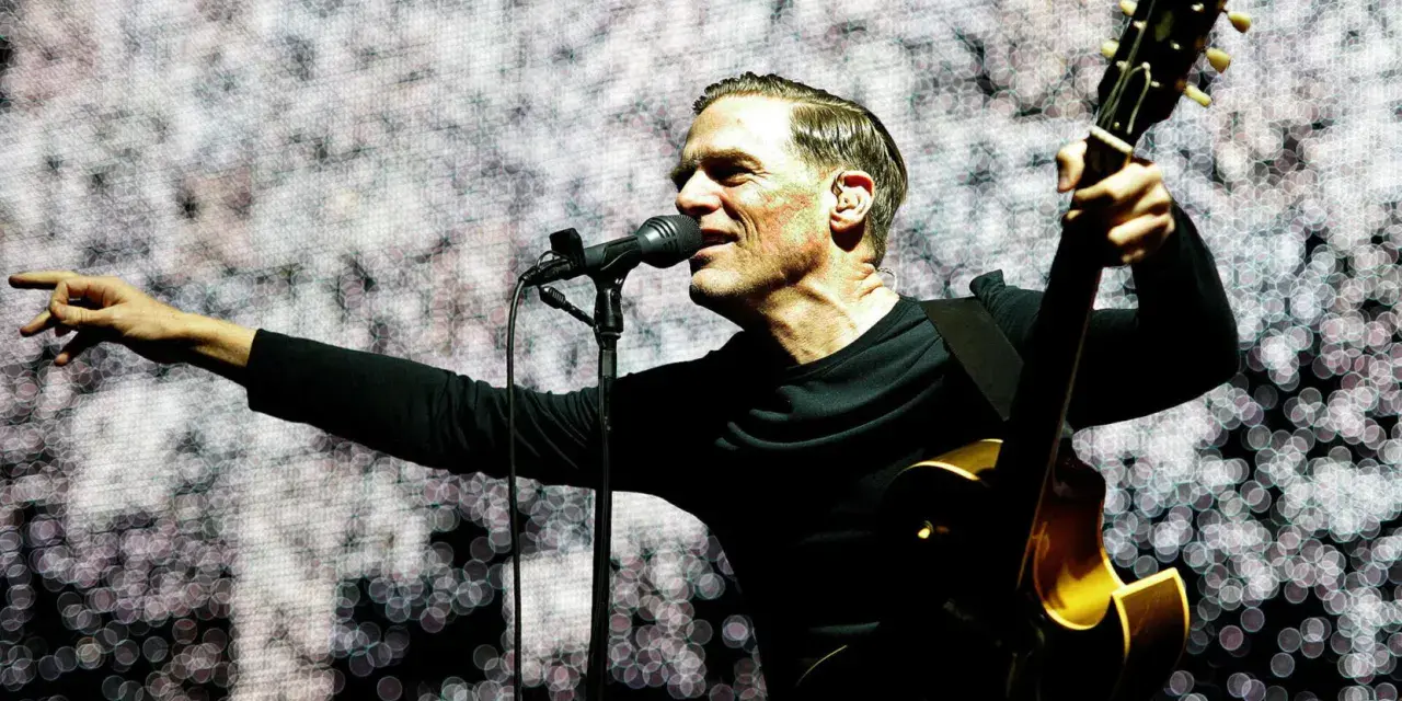 Bryan Adams na koncercie, śpiewa do mikrofonu i gra na gitarze. Wskazuje palcem na publiczność, tworząc niezapomniane chwile.