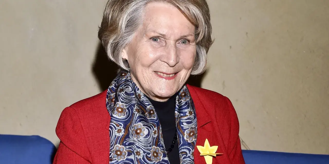 Uśmiechnięta Irena Santor w czerwonej marynarce i wzorzystym szalu, z żółtą rozetką na klapie.