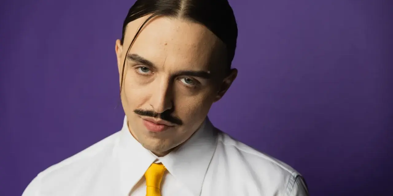 Tommy Cash w białej koszuli i żółtym krawacie, z charakterystycznym wąsem i przedziałkiem na środku głowy, na fioletowym tle.