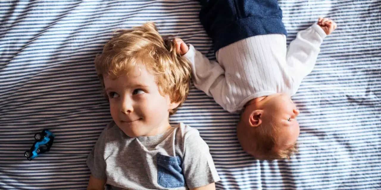 Dwóch braci, Emil i maluszek, leży na pasiastym kocyku. Emil ma blond włosy i uśmiecha się.