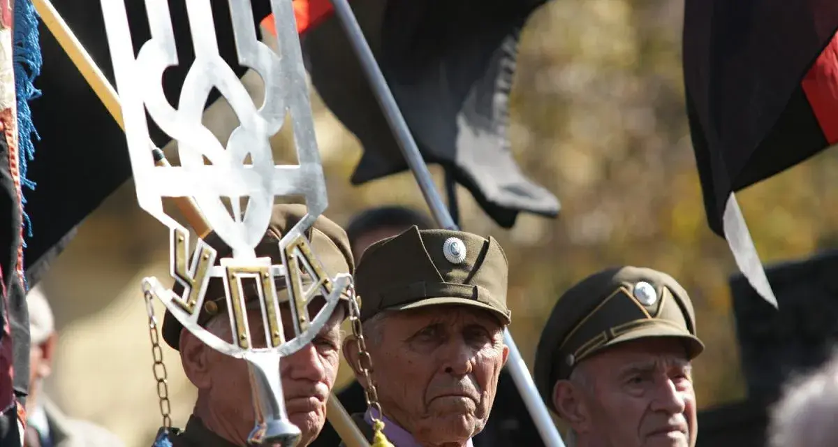 Trzech starszych mężczyzn w wojskowych czapkach, trzymających flagi i symbol "upa".
