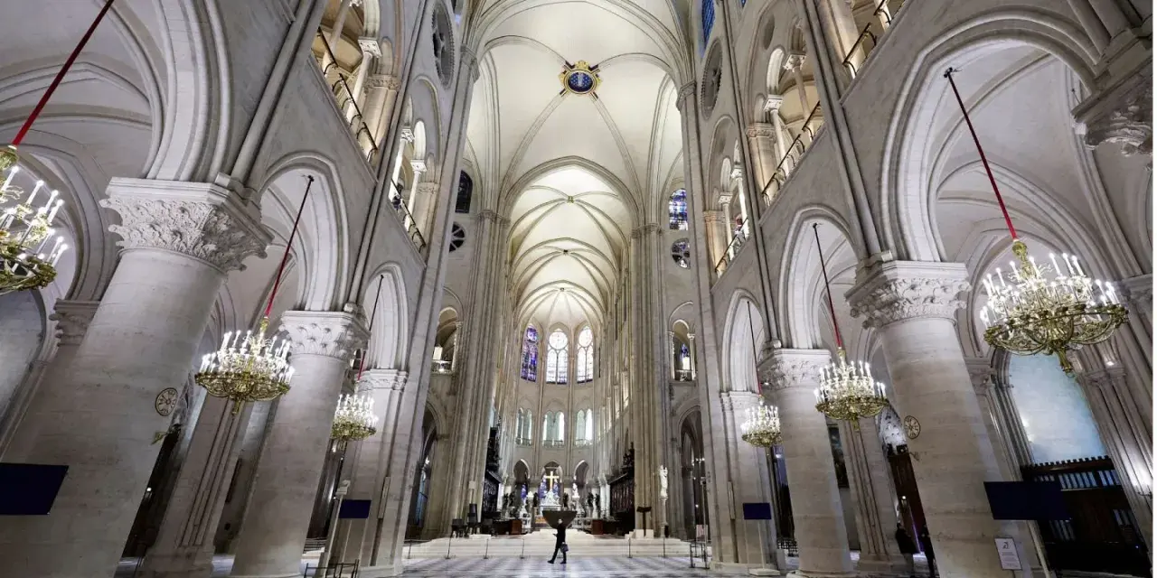 Wnętrze katedry Notre Dame z imponującymi łukami, sklepieniem i żyrandolami. Na szachownicowej posadzce widać pojedynczą postać.
