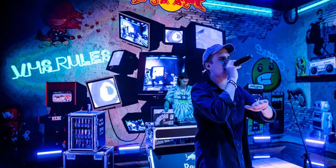 Jan rapowanie koncert w neonowym świetle. Na ścianie napisy "VHS RULES", logo Red Bulla i stare monitory.