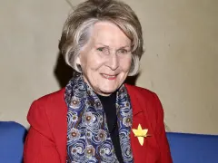 Uśmiechnięta Irena Santor w czerwonej marynarce i wzorzystym szalu, z żółtą rozetką na klapie.