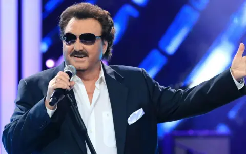 Krzysztof Krawczyk w okularach przeciwsłonecznych i granatowej marynarce śpiewa na scenie, gestykulując prawą ręką.