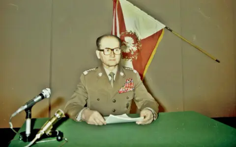 Generał w mundurze, z flagą Polski za plecami, przemawia na konferencji prasowej. To ważny moment dla Wojskowej Rady Ocalenia Narodowego.