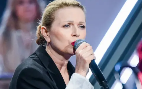 Anna Maria Jopek, w czarnej marynarce, śpiewa do mikrofonu. W tle rozmyte postacie i światła sceny.
