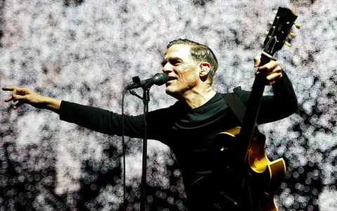 Bryan Adams na koncercie, śpiewa do mikrofonu i gra na gitarze. Wskazuje palcem na publiczność, tworząc niezapomniane chwile.