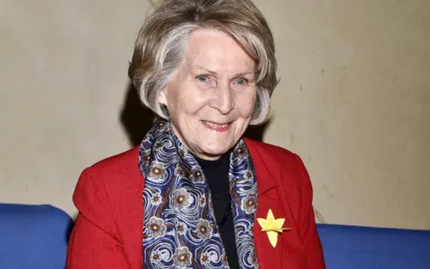 Uśmiechnięta Irena Santor w czerwonej marynarce i wzorzystym szalu, z żółtą rozetką na klapie.
