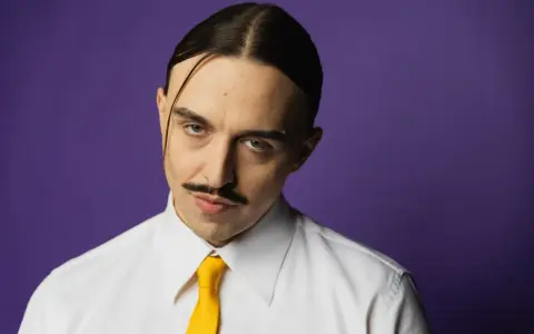 Tommy Cash w białej koszuli i żółtym krawacie, z charakterystycznym wąsem i przedziałkiem na środku głowy, na fioletowym tle.