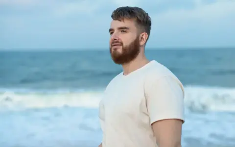 Mężczyzna z brodą na plaży, patrzący w dal. Arkadiusz - imię oznaczające "wielki władca", "wielki pan".