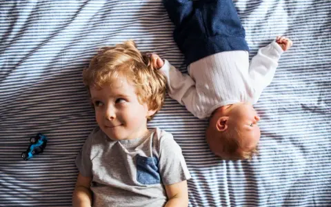Dwóch braci, Emil i maluszek, leży na pasiastym kocyku. Emil ma blond włosy i uśmiecha się.