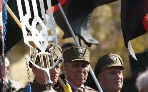 Trzech starszych mężczyzn w wojskowych czapkach, trzymających flagi i symbol "upa".