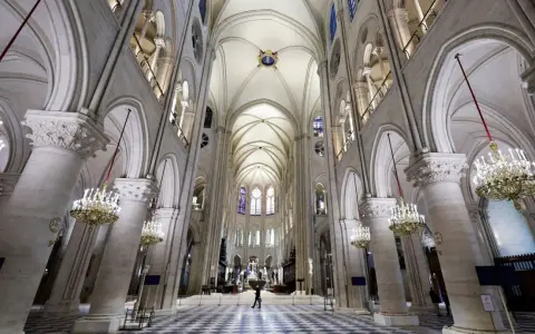 Wnętrze katedry Notre Dame z imponującymi łukami, sklepieniem i żyrandolami. Na szachownicowej posadzce widać pojedynczą postać.