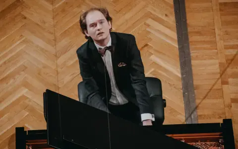Piotr Ryszard Pawlak w eleganckim garniturze pochyla się nad fortepianem, gotowy do wykonania utworu.