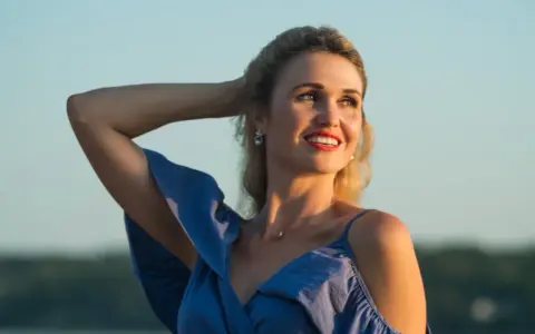 Uśmiechnięta Anita w niebieskiej sukience, z ręką we włosach. Jej imię, Anita, znaczy "łaska".