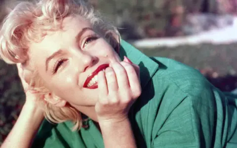 Marilyn Monroe, ikona kina, w zielonej sukience, z dłonią przy ustach, uśmiecha się promiennie.