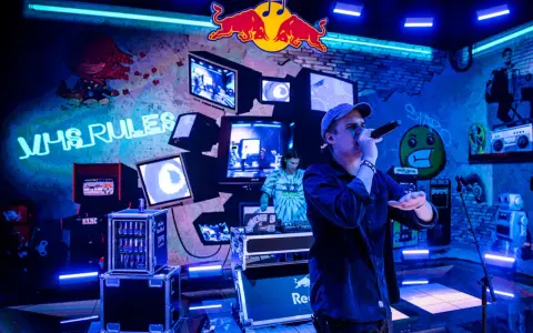 Jan rapowanie koncert w neonowym świetle. Na ścianie napisy "VHS RULES", logo Red Bulla i stare monitory.