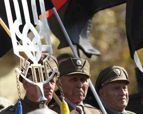 Trzech starszych mężczyzn w wojskowych czapkach, trzymających flagi i symbol "upa".
