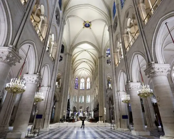Wnętrze katedry Notre Dame z imponującymi łukami, sklepieniem i żyrandolami. Na szachownicowej posadzce widać pojedynczą postać.