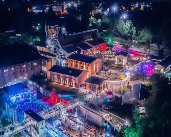 Nocny festiwal Carbon Silesia rozświetla postindustrialny krajobraz. Tłumy bawią się przy scenie, a kolorowe światła odbijają się od budynków dawnej kopalni.