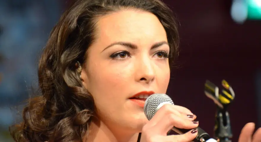 Caro Emerald śpiewa do mikrofonu, zbliżenie na twarz i dłonie.