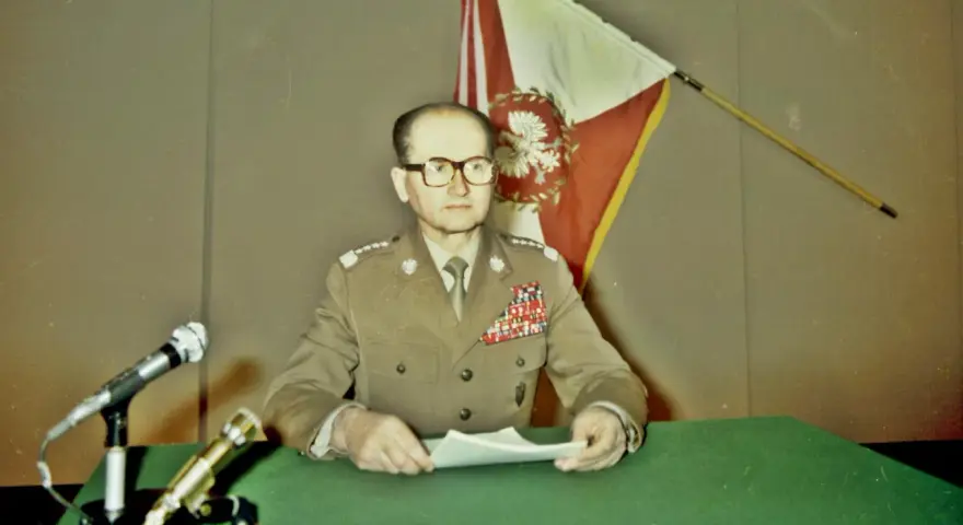 Generał w mundurze, z flagą Polski za plecami, przemawia na konferencji prasowej. To ważny moment dla Wojskowej Rady Ocalenia Narodowego.