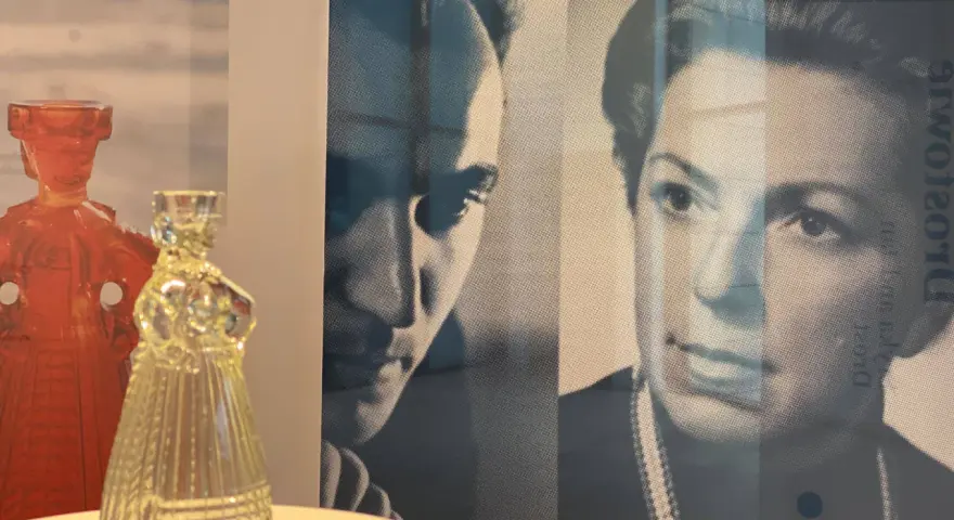Szklana figura w kształcie sukni obok czerwonej postaci. W tle portret Eryki Trzewik Drost.