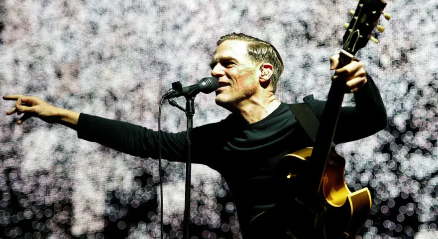 Bryan Adams na koncercie, śpiewa do mikrofonu i gra na gitarze. Wskazuje palcem na publiczność, tworząc niezapomniane chwile.