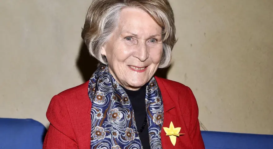 Uśmiechnięta Irena Santor w czerwonej marynarce i wzorzystym szalu, z żółtą rozetką na klapie.