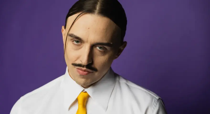 Tommy Cash w białej koszuli i żółtym krawacie, z charakterystycznym wąsem i przedziałkiem na środku głowy, na fioletowym tle.