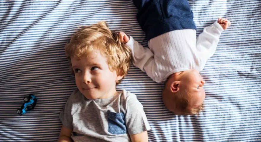 Dwóch braci, Emil i maluszek, leży na pasiastym kocyku. Emil ma blond włosy i uśmiecha się.