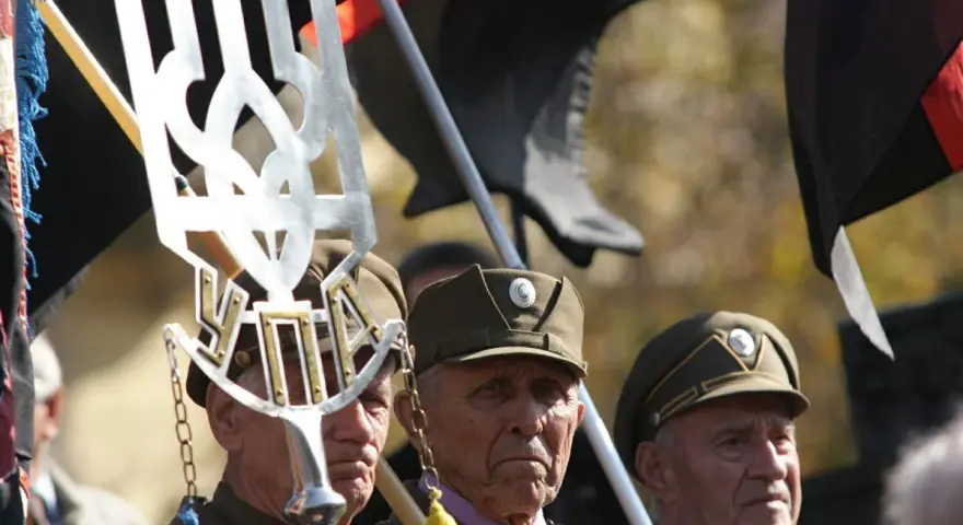 Trzech starszych mężczyzn w wojskowych czapkach, trzymających flagi i symbol "upa".