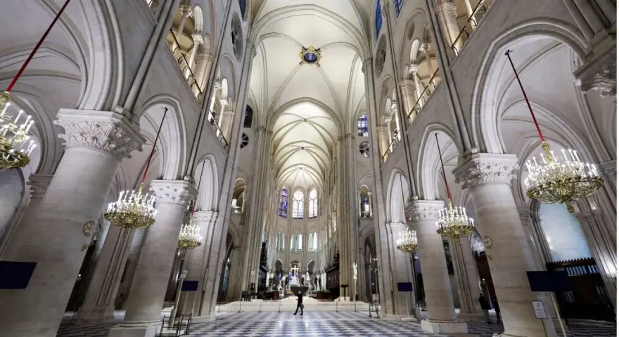 Wnętrze katedry Notre Dame z imponującymi łukami, sklepieniem i żyrandolami. Na szachownicowej posadzce widać pojedynczą postać.