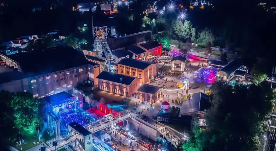 Nocny festiwal Carbon Silesia rozświetla postindustrialny krajobraz. Tłumy bawią się przy scenie, a kolorowe światła odbijają się od budynków dawnej kopalni.