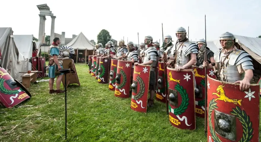 Rzymscy legioniści w pełnym uzbrojeniu, z tarczami i włóczniami, gotowi do bitwy. W tle namioty i ruiny.