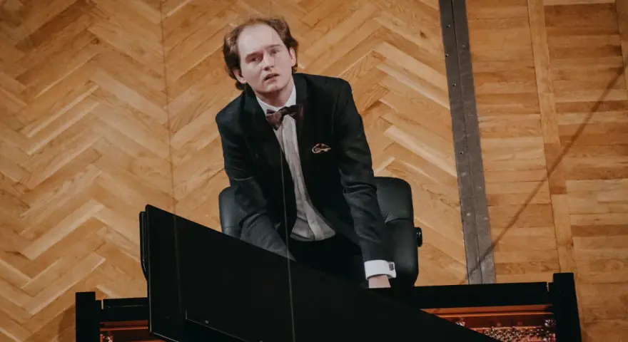 Piotr Ryszard Pawlak w eleganckim garniturze pochyla się nad fortepianem, gotowy do wykonania utworu.