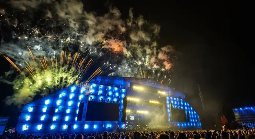 Nocny koncert z fajerwerkami nad sceną. Tłum ludzi podziwia widowisko, a nad nimi rozbłyskują kolorowe petardy.