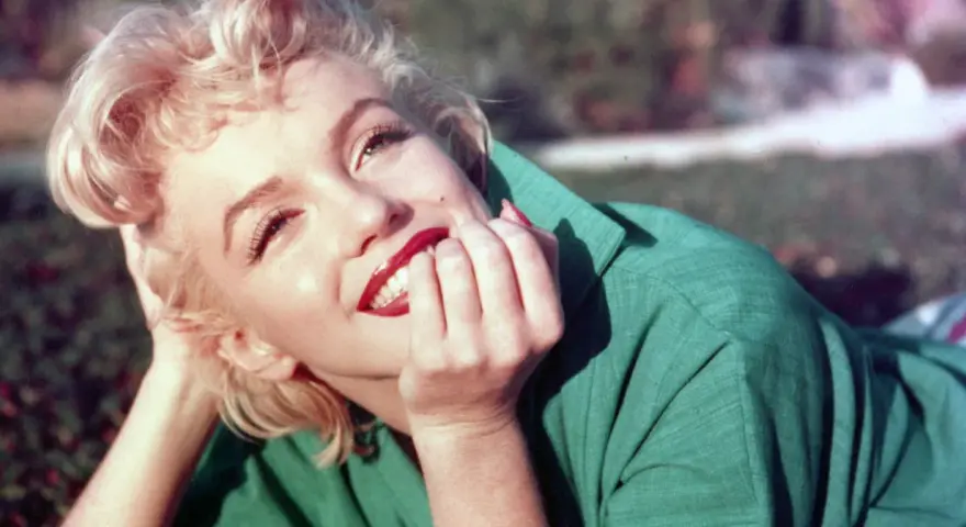 Marilyn Monroe, ikona kina, w zielonej sukience, z dłonią przy ustach, uśmiecha się promiennie.